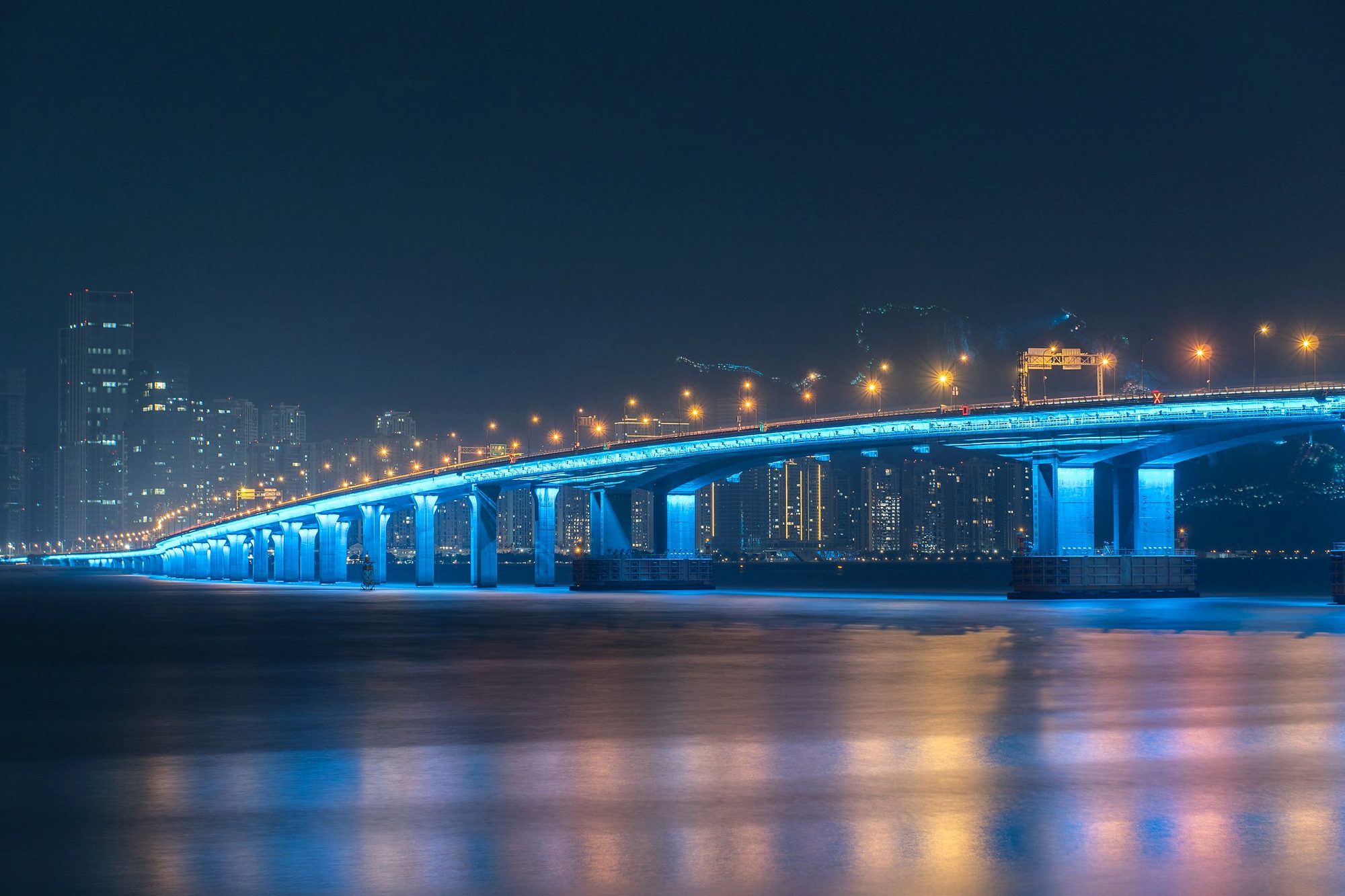 Pont illuminé en bleu au-dessus d’un fleuve, la nuit, devant une ville